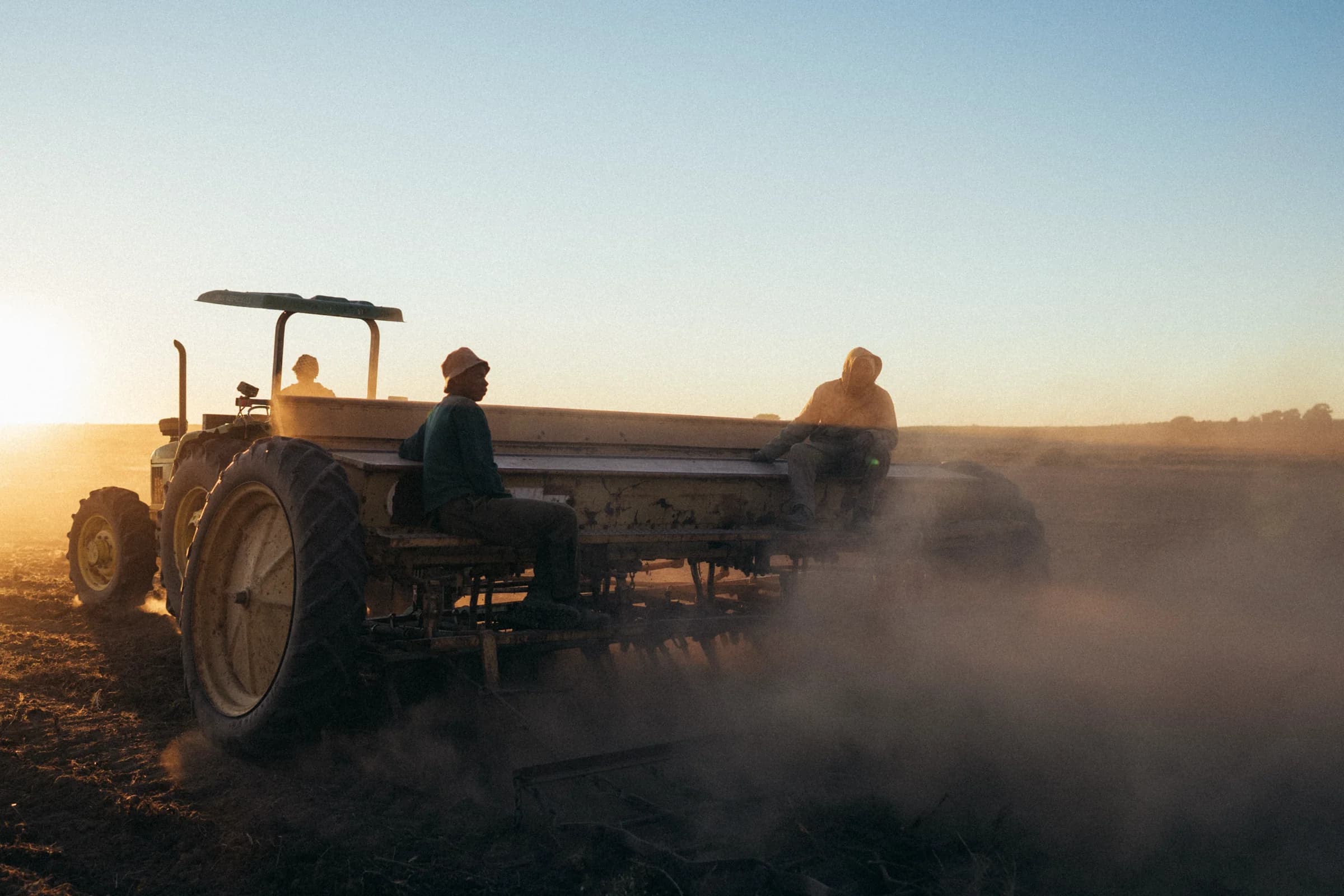 A tractor pulling a planter across a dusty land at sunset, two people aboard
