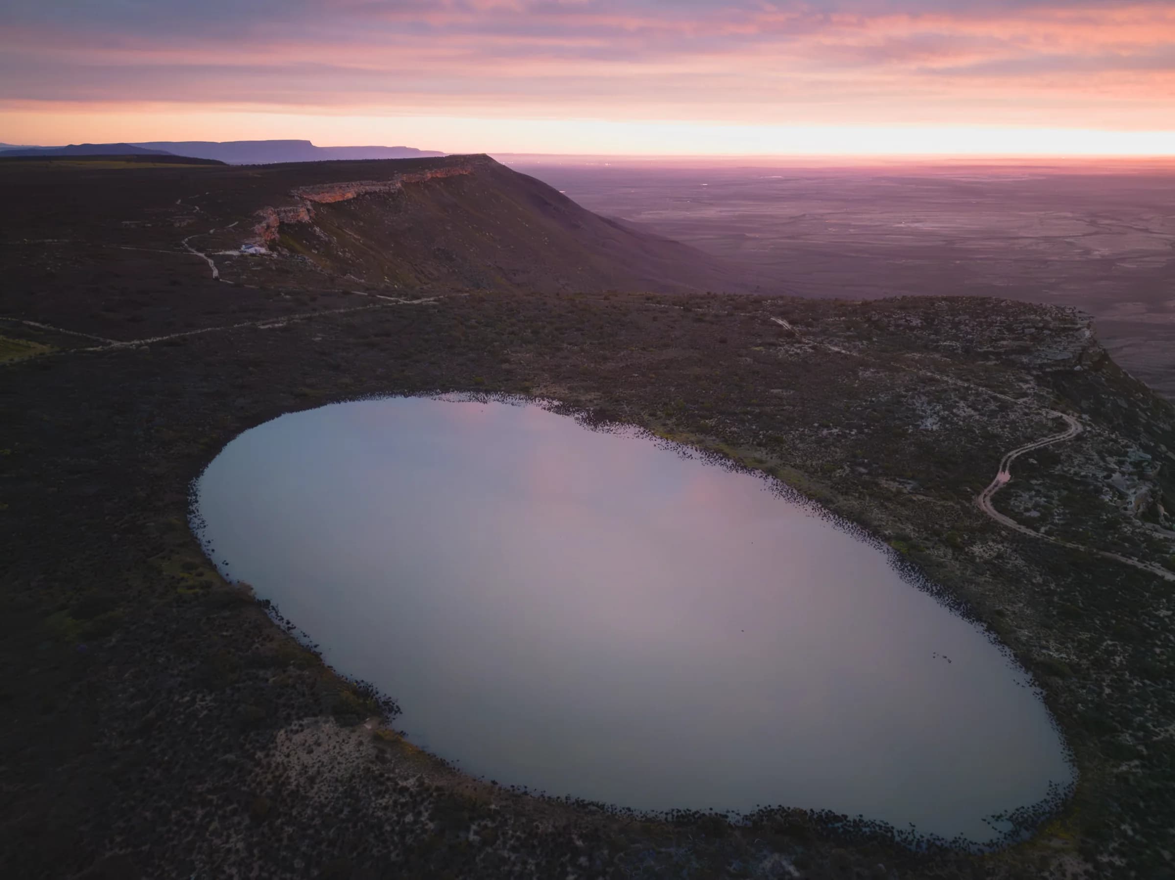 An aerial view of an oval dam on the edge of the krans at sunset, the Knersvlakte beyond